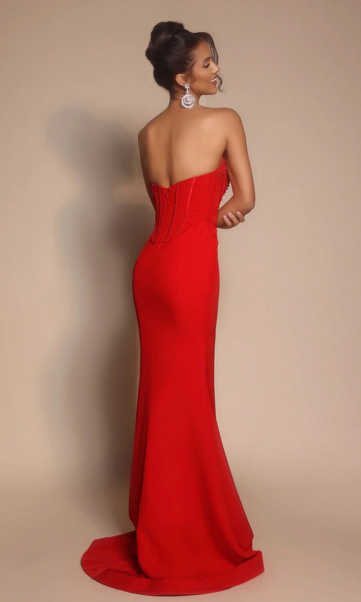 A woman stands with her back to the camera against a plain beige background, wearing the Portia and Scarlett PS26008 long formal strapless evening gown featuring a beaded corset.